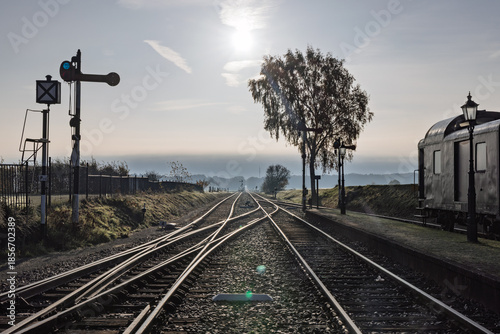 Railway tracks diverging at a switch with old signals and a vintage carriage, silhouetted against a bright backlit sun and misty autumn sky.
