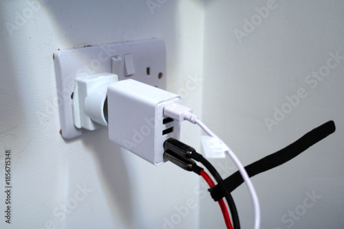 A white electrical adapter is plugged into a wall socket, connected through a power converter. Three USB ports on the adapter are in use with attached cables. The wall plate shows a UK-style socket.