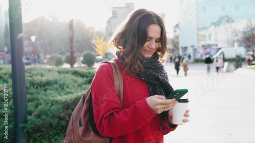 A pretty young woman uses her mobile phone while walking through the streets of a European city on a warm autumn day.