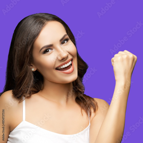 Image of happy gesturing beautiful woman with open mouth raising hand up with clenching fist, wear white casual clothing, isolated violet purple background. Expressive emotional brunette girl. Square