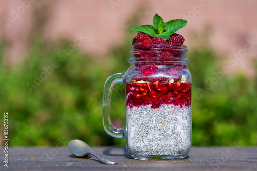 Healthy chia pudding in a glass mug with fresh red raspberry, chia seeds, jam, walnuts, coconut milk and honey for breakfast on a wooden table on nature background, closeup
