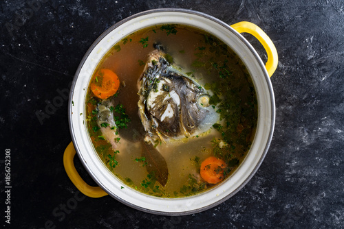 Healthy soup with carp fish head and fillet with vegetables in a saucepan on the table, closeup, top view