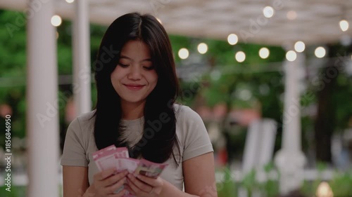 Young woman at home excitedly counting cash rupiah in hands, smiling and celebrating unexpected financial gain. Asian girl happy about lottery win, salary or surprise money profit.