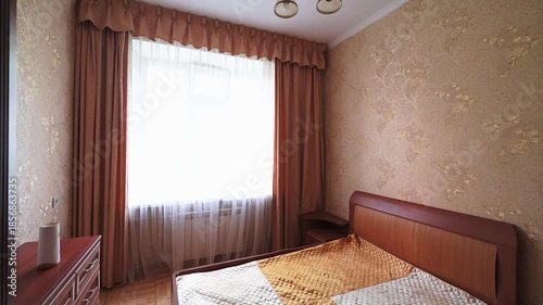 Cozy bedroom with wooden bed, patterned wallpaper, large window with sheer curtains, wooden wardrobe, ceiling lamp, warm beige tones, tidy layout