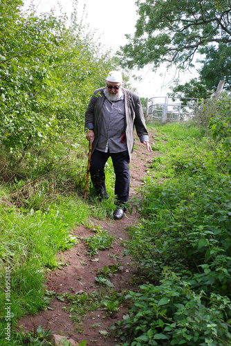 Senior man walking on a narrow dirt path in Wick Golden Valley Nature Reserve. He is leaning on a wooden cane. The path is lined with bushes and trees, leading toward a gate.