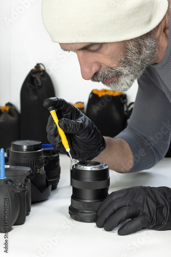 A bearded man wearing gloves carefully repairs a camera lens with a small screwdriver, surrounded by camera equipment on a clean worktable.