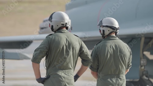 Rear view of two pilots in flight gear observing a fighter aircraft during ground operation on an airbase