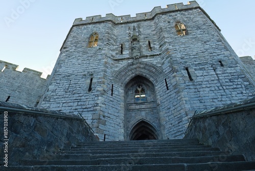 Imposing grey stone castle gatehouse rises, viewed low-angle, steps leading up; crenellations, arched entrance, dim light, medieval, formidable, symmetrical, textured stone, solemn mood in Caernarfon 