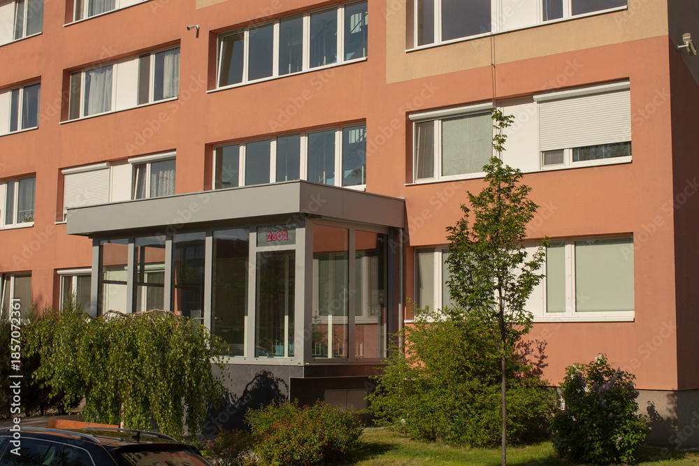 Fototapeta premium Glass entrance with warm coral facade and visible address, landscaped front with shrubs and small tree, reflective doors and sunny