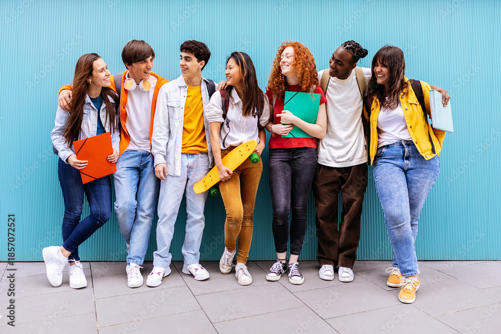 Fototapeta premium Young happy multiracial student friends standing over isolated blue background. Diverse teenage people having fun talking leaning on campus building college wall. Education and youth concept
