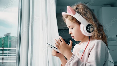 Young child using smartphone while wearing headphones at home near window