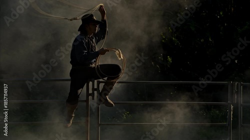 A cowboy sitting on a corral fence skillfully spinning a lasso in dusty morning light