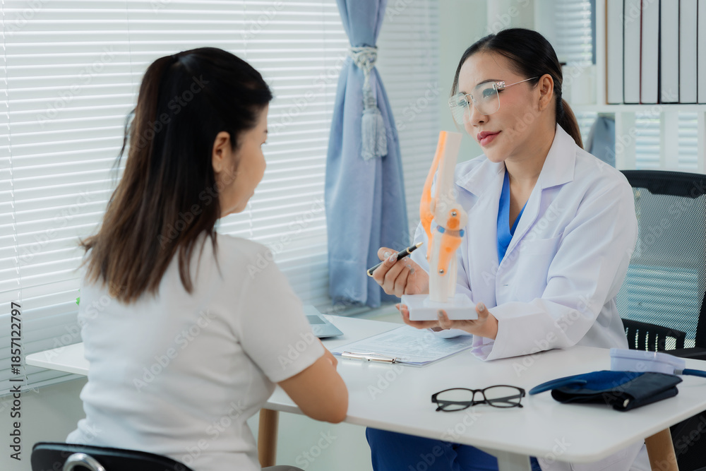 Fototapeta premium An Asian female doctor and her patient were discussing bones, and she pointed a pen at an anatomical model of a human knee joint placed on a table.