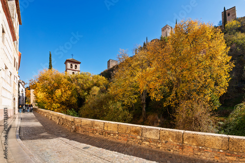 The Alhambra rises above the historic Carrera del Darro street along the Darro River in the old town of Granada, Spain at Autumn with the Iglesia de San Pedro y San Pablo church behind.