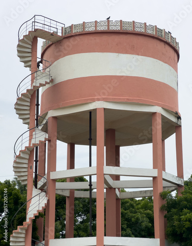 Elevated water storage tank with blue railings on stilts, standing against green trees and cloudy sky, a common rural water supply infrastructure.