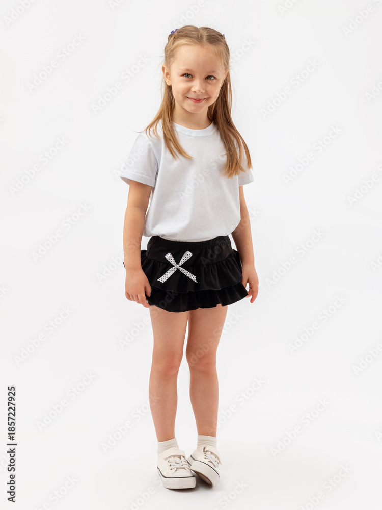 Fototapeta premium A modest Caucasian girl with a kind smile and slender legs is standing on a white background. She is wearing a T-shirt, a skirt, and sneakers.