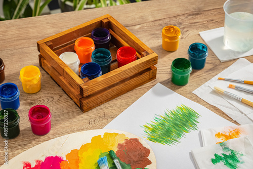 Artist's workplace. On the table, there is a wooden box filled with paints of different colors and a glass of water. A drawing and paintbrushes. Top view