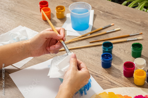 Women's hands wipe the brush with a paper towel to remove excess water after painting. There are different colors of gouache and brushes on the table. Lifestyle. Close-up. Side view.