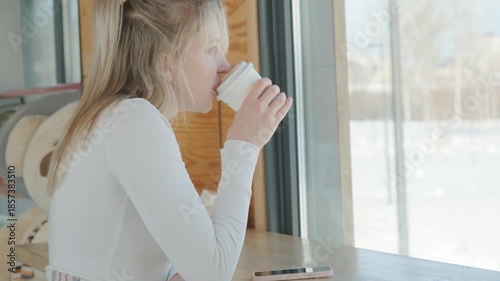 The caucasian young woman is drinking coffee in a coffee shop in winter in front of a window