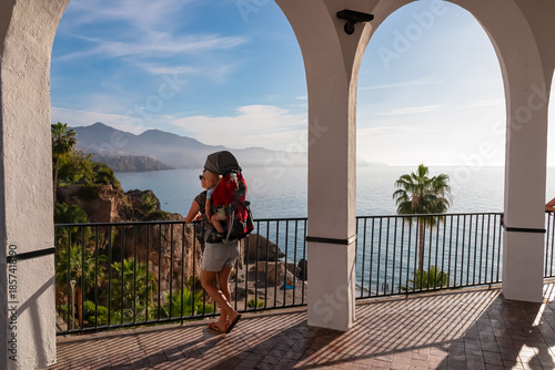 A mother carrying her baby in a backpack stands beneath the white arches of Balcon de Europa, admiring the panoramic coastal cliffs and sea views in the sunny destination of Nerja in southern Spain.