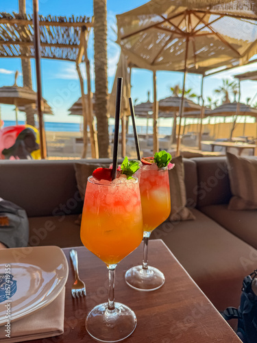 Two vibrant fruit cocktails with black straws resting on a wooden table under thatched umbrellas and palm trees at a sunny beach bar in Marbella, invoking a relaxing holiday vibe on the Mediterranean.