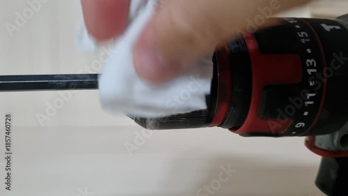 Close up view of a hand cleaning the chuck and bit of a red cordless power drill with a wet wipe for tool maintenance and hygiene