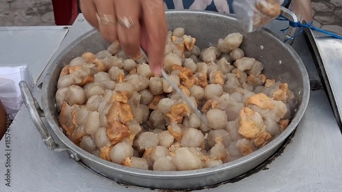 Street food vendor serving siomay and cilok, stirring hot dumplings while assisting customers at a local stall. Authentic Indonesian street food scene with fresh, homemade snacks.