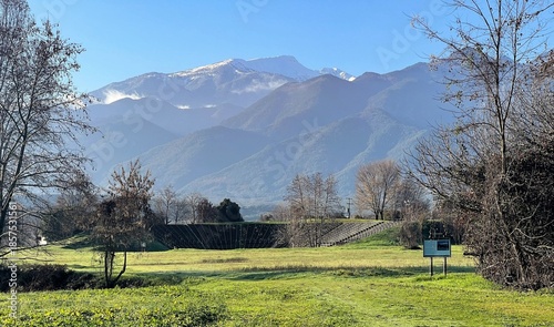 Photo of Mount Olympus rising above the archaeological site of Dion and its Hellenistic Theatre,framed by winter trees, a sunlit green meadow and a clear blue sky.