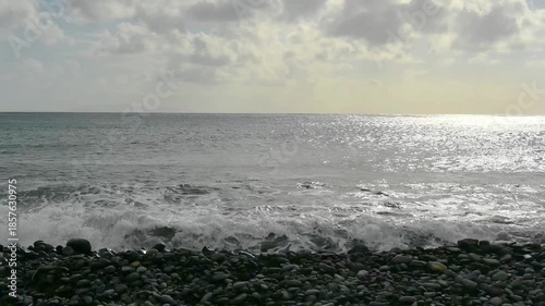 waves of the Atlantic Ocean with reflection of sunlight splashing on grey volcanic rocks and pebbles on a beach 