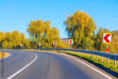 Winding asphalt road curves right through golden autumn trees under blue sky, lined with metal guardrails. Chevron signs mark the bend amid lush yellow foliage and distant hills