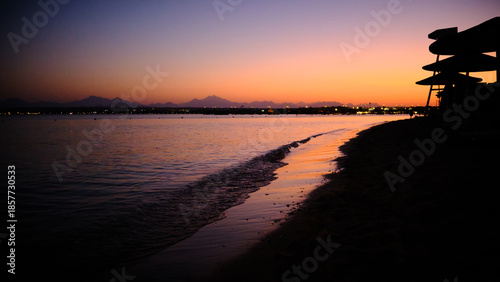 Calm seaside shoreline at sunset with gentle waves washing onto the beach. Warm orange and purple tones fill the sky, creating tranquil coastal atmosphere with silhouettes of distant landforms