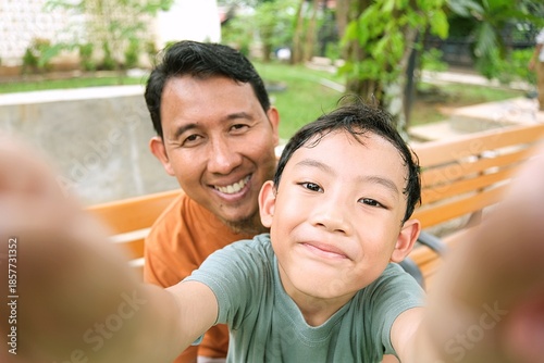 Happy Asian father and young son taking a selfie together outdoors, enjoying quality family time in a green park, representing wellbeing, sustainability, and future generations.