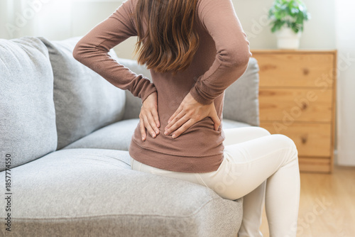 Fotografie Pain body muscles stiff problem, suffer asian young woman painful back, symptom injury ache from long study work, hand holding massaging or rubbing lower back hurt, sore sitting on sofa at home