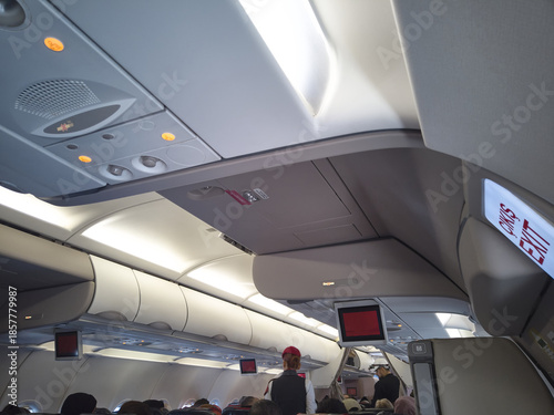 Interior view of commercial airplane cabin with overhead compartments, ceiling lighting, information screens, and seated passengers