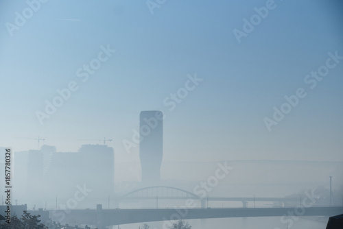 Atmospheric urban landscape showing city skyline partially obscured by dense fog, with bridge crossing river in foreground