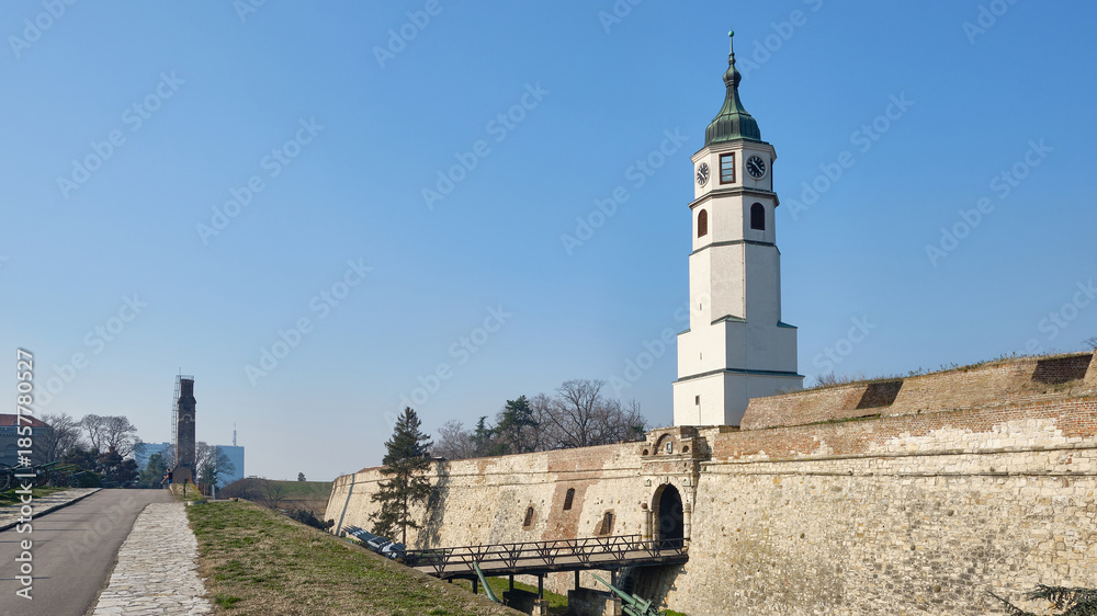 Obraz premium View of historic fortress with tall clock tower and massive stone walls under clear blue sky. Sahat Tower of Kalemegdan fortress, Belgrade, Serbia