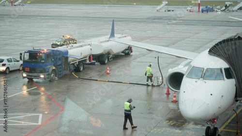 Commercial aircraft refueling process at airport apron on rainy day. Fuel tanker truck pumps kerosene into jet wing while ground crew in safety vests monitor logistics near terminal gate.