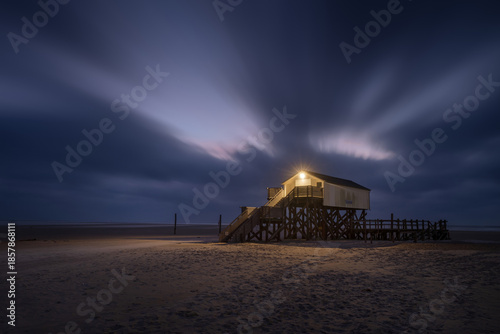 Illuminated Stilt House on Sandy Beach at Night with Dramatic Long Exposure Sky and Streaking Clouds.