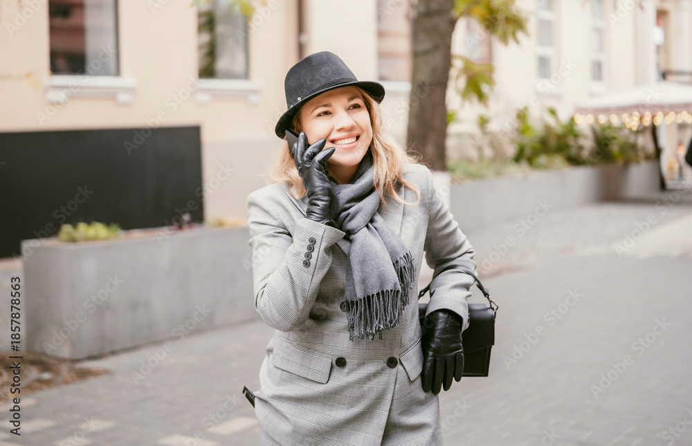 Fototapeta premium Business woman in grey suit, hat, scarf and gloves talking on smartphone while walking through autumn city after business lunch. Lifestyle, communication, business street and city life concept.