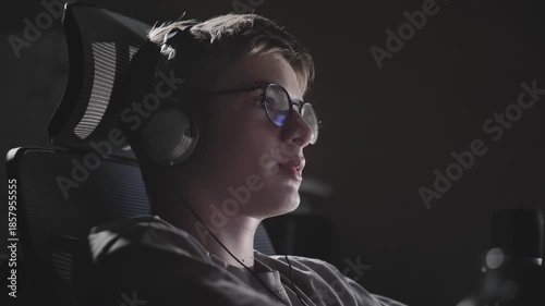 Handheld cinematic shot of teenage gamer boy with headset microphone playing online game and communicating with teammates, dramatic backlighting