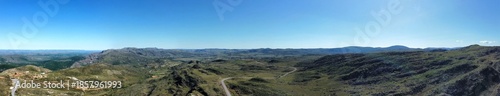 Wide panoramic aerial view of Serra do Cipó, Minas Gerais, Brazil, featuring vast mountain landscapes, winding roads, native vegetation, and a clear blue sky