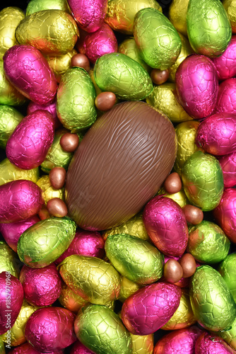 Pile or group of multi colored and different sizes of colourful foil wrapped chocolate easter eggs in pink, red, yellow and lime green with a large brown milk chocolate egg in the middle.