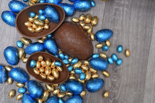 Pile of colourful foil wrapped chocolate easter eggs in blue, and gold. Large halves of a brown milk chocolate egg have mini eggs inside, on a grey wooden background.