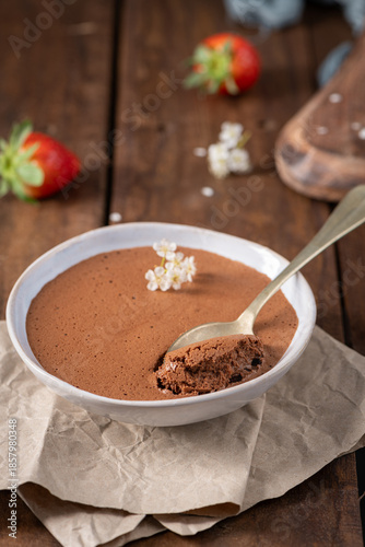 Velvety chocolate mousse in a white bowl, a golden spoon taking a bite, adorned with delicate white flowers, resting on crumpled parchment paper and a rustic wooden surface