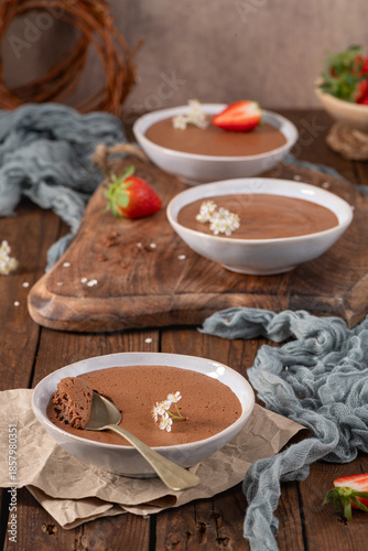 Individual servings of chocolate mousse in white bowls, garnished with strawberries and tiny white flowers, arranged on a rustic wooden board and table with soft fabric