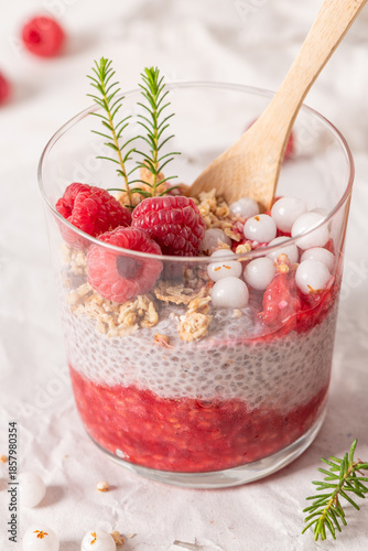 A single glass of layered raspberry and chia seed pudding topped with fresh raspberries and white crowberry on a rustic white table