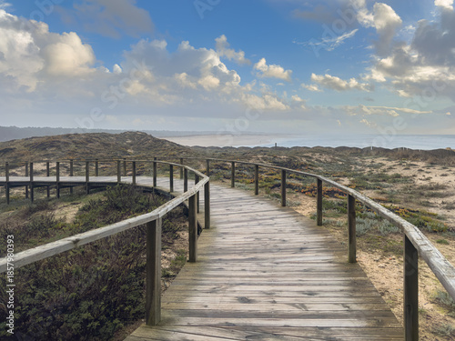 A wooden boardwalk curves across sandy coastal dunes covered with low vegetation, offering a panoramic view of the Furadouro beach and Atlantic Ocean under a bright, cloudy sky in Ovar Portugal