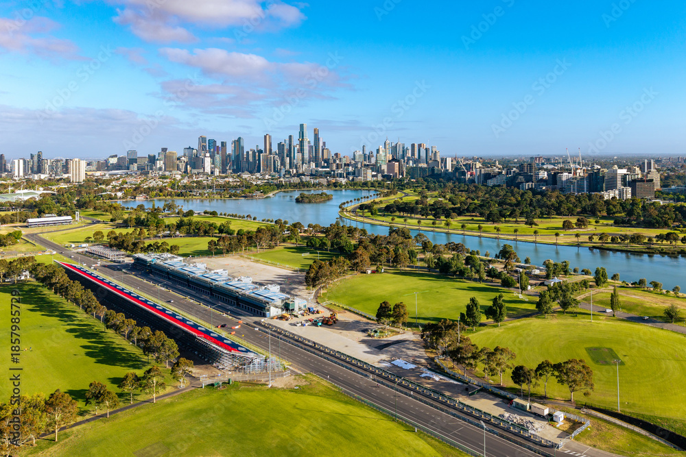 Obraz premium Elevated view of Melbourne's striking city skyline, Albert Park Lake, and the race circuit with grandstands under a bright blue sky.
