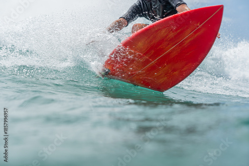 A young man in a black wetsuit rides a wave on a red surfboard and carves a hard turn in front of the camera.