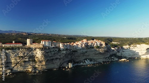 Aerial view of Bonifacio perched on dramatic white limestone cliffs above the Mediterranean Sea in Corsica, France. Picturesque historic buildings overlook the rugged coastline and clear blue water.